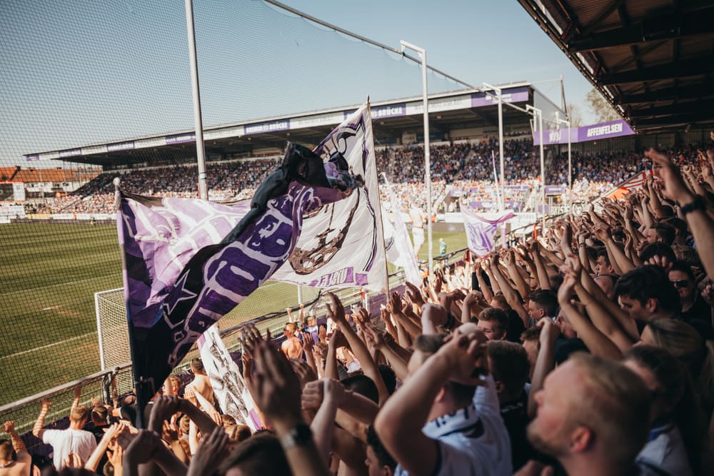erfolgsgeschichte-vfl-2 Eine Foto von jubelnden VfL Fans beim Zuschauen eines Spiels.
