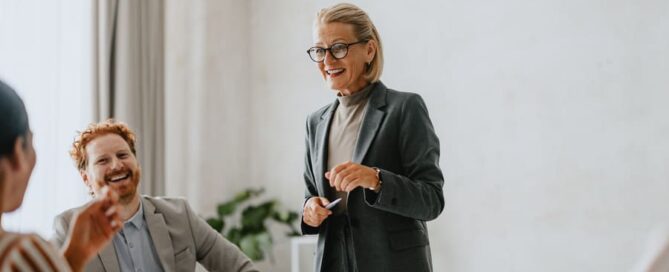 Eine Frau steht im Blazer vor einer Gruppe Mitarbeitenden