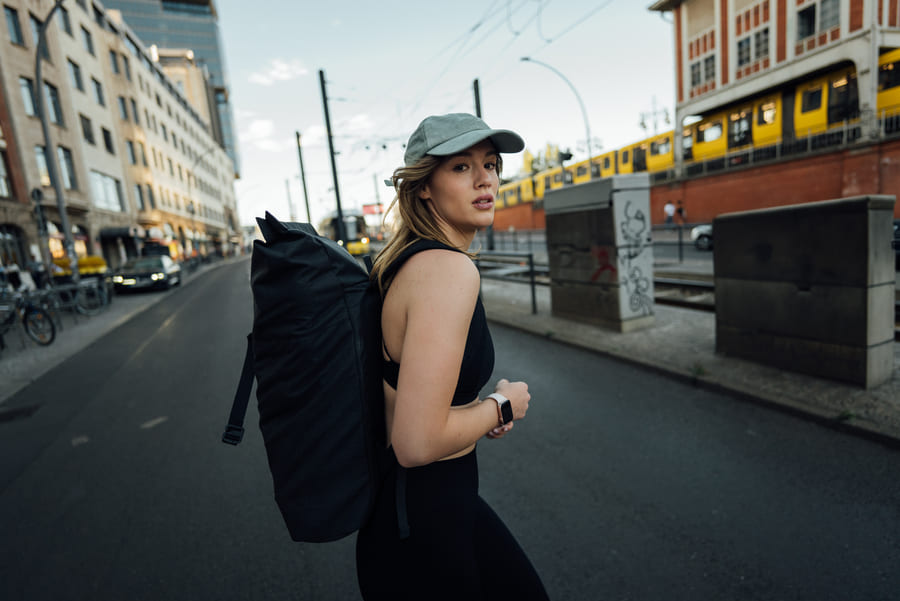 Eine Frau mit einer Cap und Rucksack auf überquert eine Straße und schaut in die Kamera.