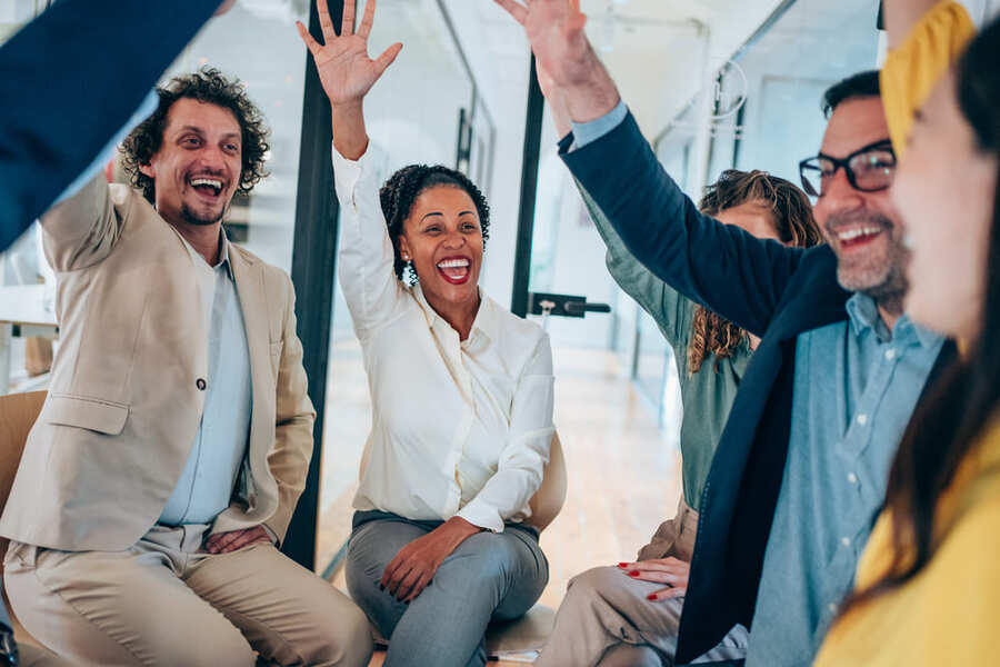 Kosten-Firmenfitness Eine Gruppe Mitarbeitende sitzen im Büro und schmeißen die Hände in die Luft und freuen sich.